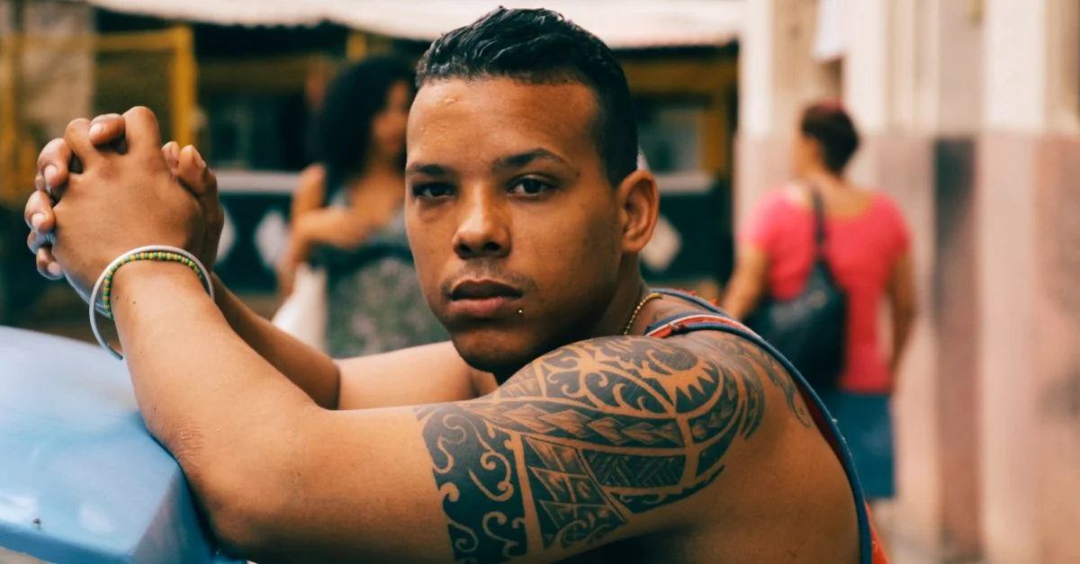 Retrato de un hombre apoyado de manera casual, con un tatuaje tribal visible, al aire libre durante el día.
